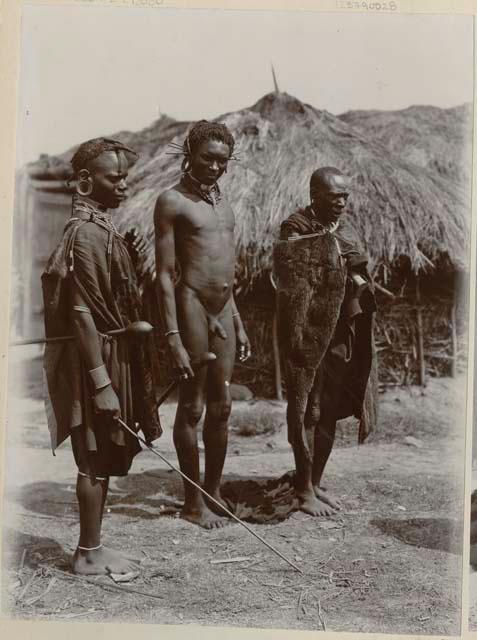 Three men standing, with huts in background