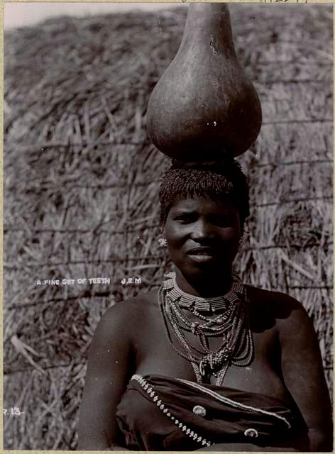 Woman with pot or gourd on her head, in front of a hut