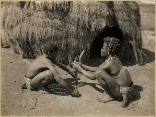 Man and boy procuring fire in front of a hut