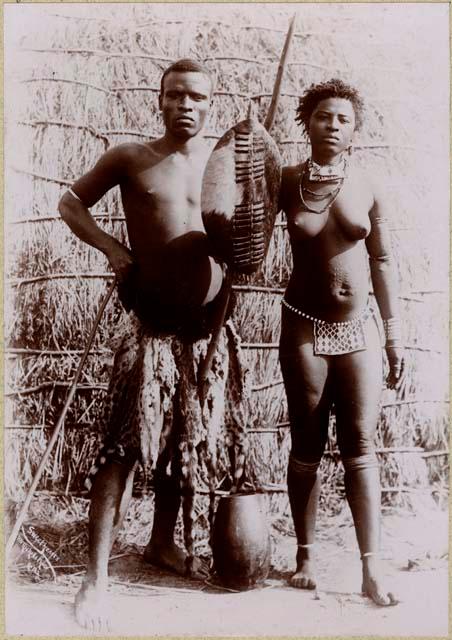 Man and woman standing in front of a hut with spear and shield