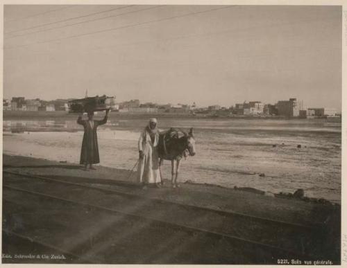 Man holding a trap and woman with donkey, with Suez in background