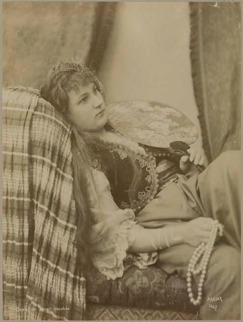 Studio portrait of a woman reclining on a couch with a fan and holding pearls