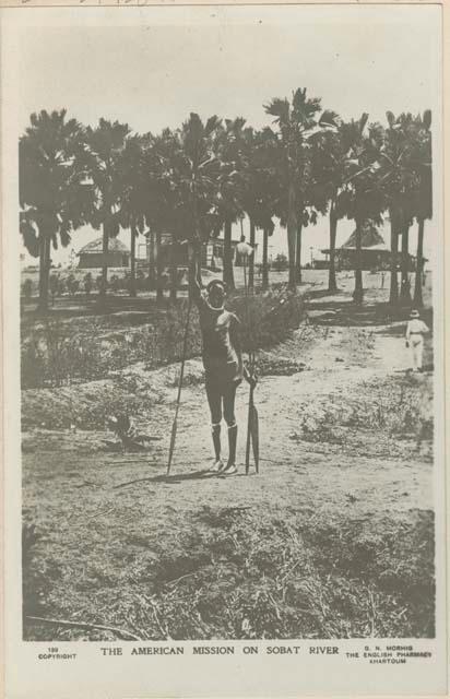 Man standing and holding a spear, trees and huts in background