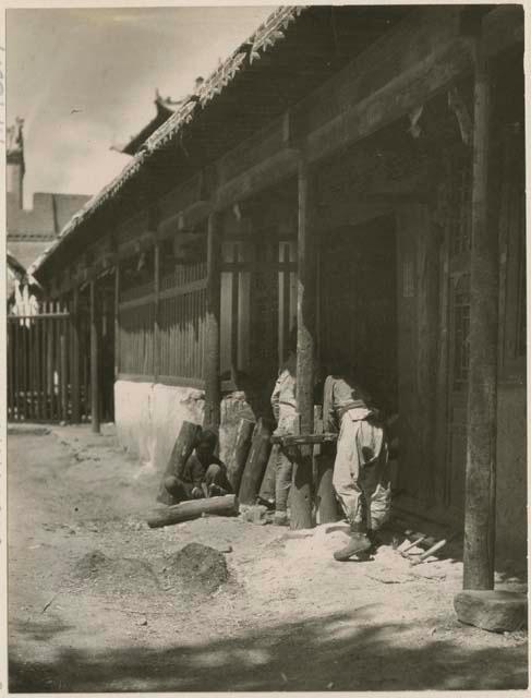 Boys sawing logs in front of carpenter shop