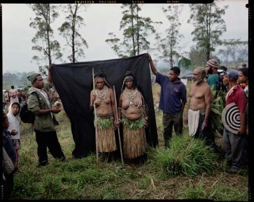 Color image: two women Sing-Sing performers with black headdresses, staffs and plant fiber skirts