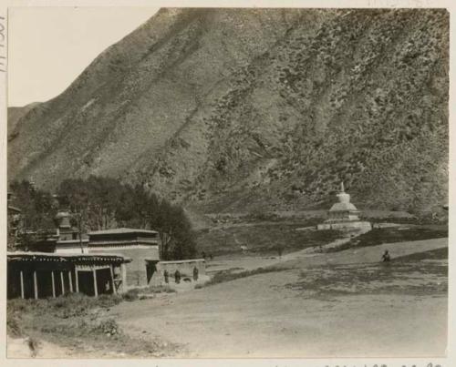 White stupa and sheds with prayer wheels among the hills, with people visible