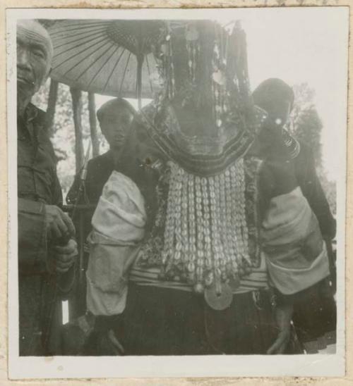 Collar of cowries, coins, and bells, possibly being worn by a woman, with people in the background