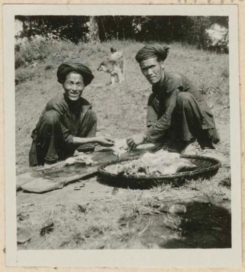 Two men sitting outside cutting meat