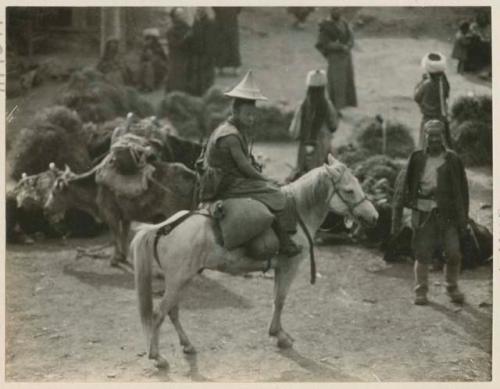 Lama sitting on horse and wearing pointed hat, leaving Labrang, Tibet, with people in background