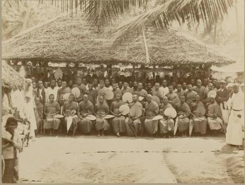 Group of men standing and seated by large thatched structure