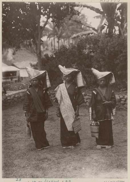 Three women in traditional dress outside, carrying bags and with hats