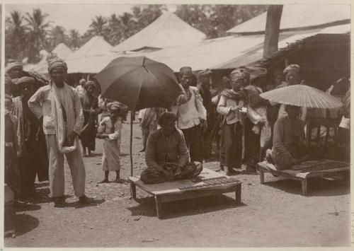 Two men sitting on small platforms with umbrellas, with objects, possibly coins, stacked in front of them.  Possibly part of an outdoor market, with people and awnings in the background.