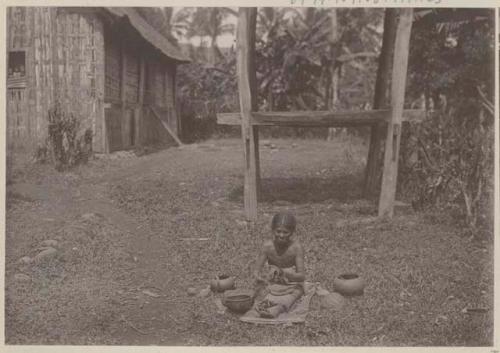 Potter sitting on the ground outside working with tools, pots, and materials