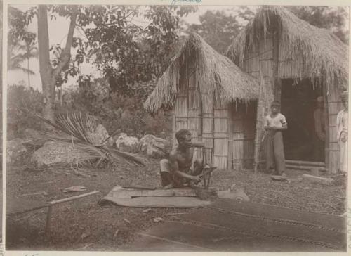 Man kneeling outside using machete, people and thatched structures in background