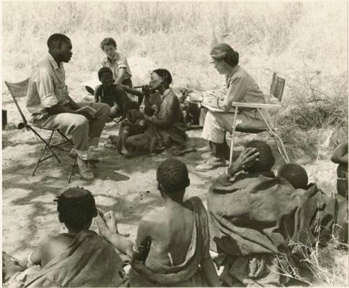 Group of people including Lorna Marshall and Kernel Ledimo seated in chairs, !U and !Ungka Norna seated between them, Elizabeth Marshall Thomas in the background, and other unidentified people