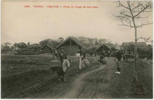 Women carrying large bundles along a path, buildings in background