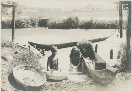 Two children on riverbank beside canoes