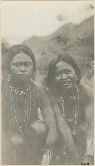 Two women wearing necklaces of beads, brass and shell, sitting