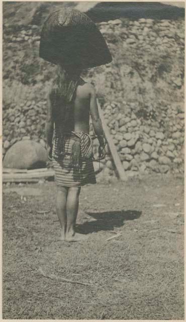 Ma-'gwė balancing a  tu-'auñ (combination rain-shield and carrying basket) on her head, view from behind