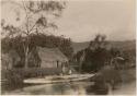 People on double canoe in river, next to shore with thatched structures