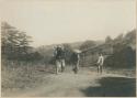 Two women carrying goods over the trail, with man standing at right