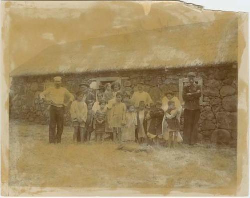 Group of people and children standing outside stone house with thatched roof