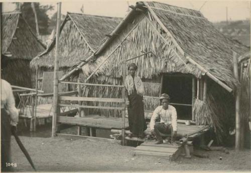 Moro man and woman on porch of thatch-roofed house