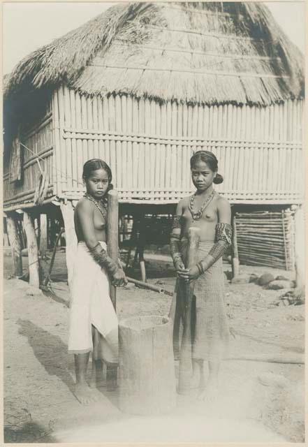 Two Tinguian girls standing by a rice mortar in front of house