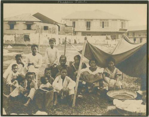 Group sitting under cloth tent, buildings