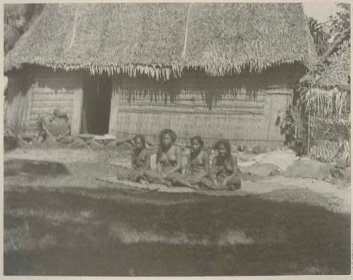 Four women sitting on the ground in front of thatched structure