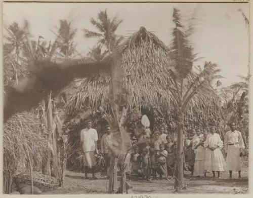 Group of adults and children in front of large thatched structure