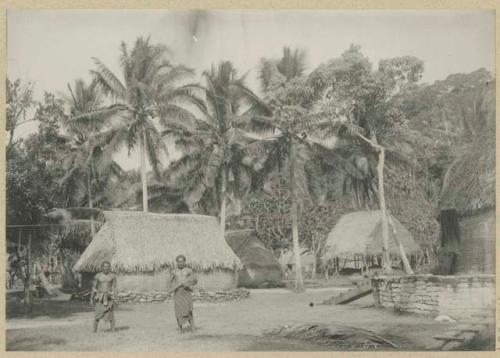 Two men in front of village structures and palms