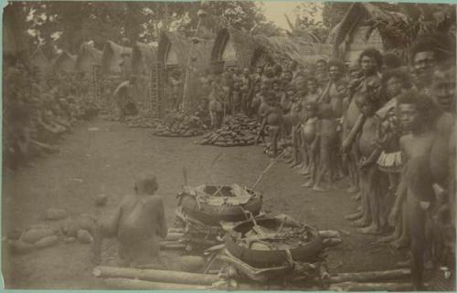 People gathered in front of row of huts, man seated by baskets on logs