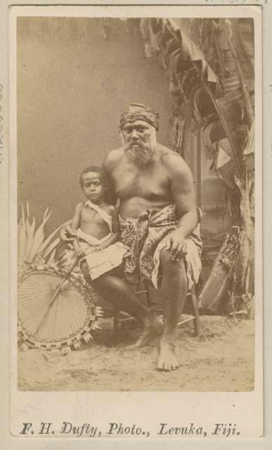 Posed studio portrait of Ratu Timothy with a child and fan