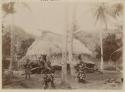 Men outside thatched structure, holding implements