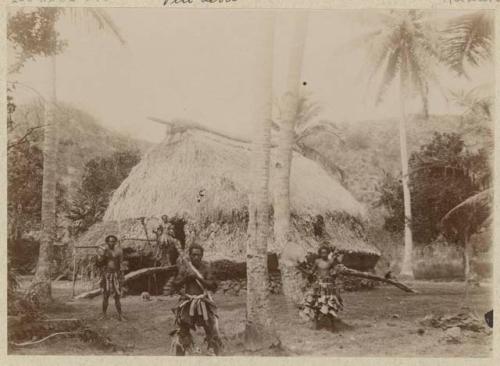 Men outside thatched structure, holding implements