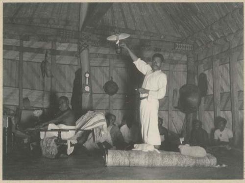 Ratu Epeli [Nailatikau I] in chair and Ratu [Penaia] Kadavulevu standing at light, in Ratu Epeli's house, with men sitting in the background