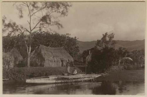 Two people in double canoe, in front of shore with thatched structures