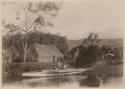 Two people with double canoe, in front of shore with thatched structures