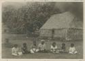 Woman seated on ground in front of thatched building and tree