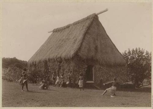 Thatched chief's house with people in front of it, including men with weapons