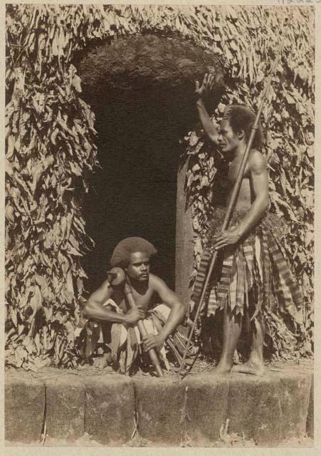 Two men at doorway, one sitting with club and one standing with spear