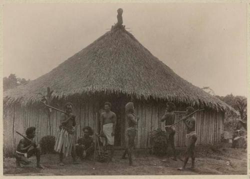 People in front of thatched house, some holding clubs