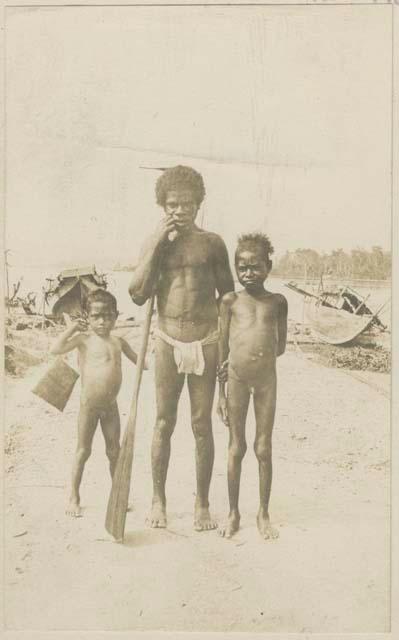 Man with oar and two children standing on shore in front of boats