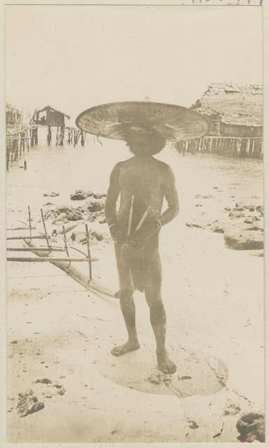Man with large hat and objects standing on shore in front of structures