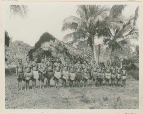 Row of women standing outside, in front of thatched structure