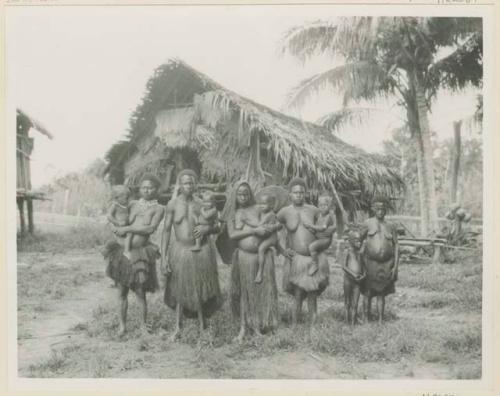 Group of women and children standing outside, in front of thatched structure