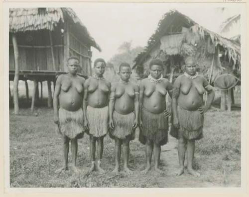 Row of women standing outside, in front of thatched structures