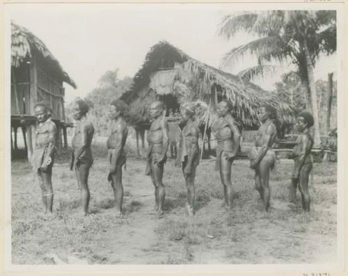 Row of men standing in profile outside, in front of thatched structures