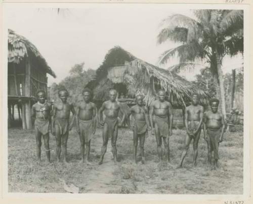 Row of men standing outside in tapa loincloths, in front of thatched structures